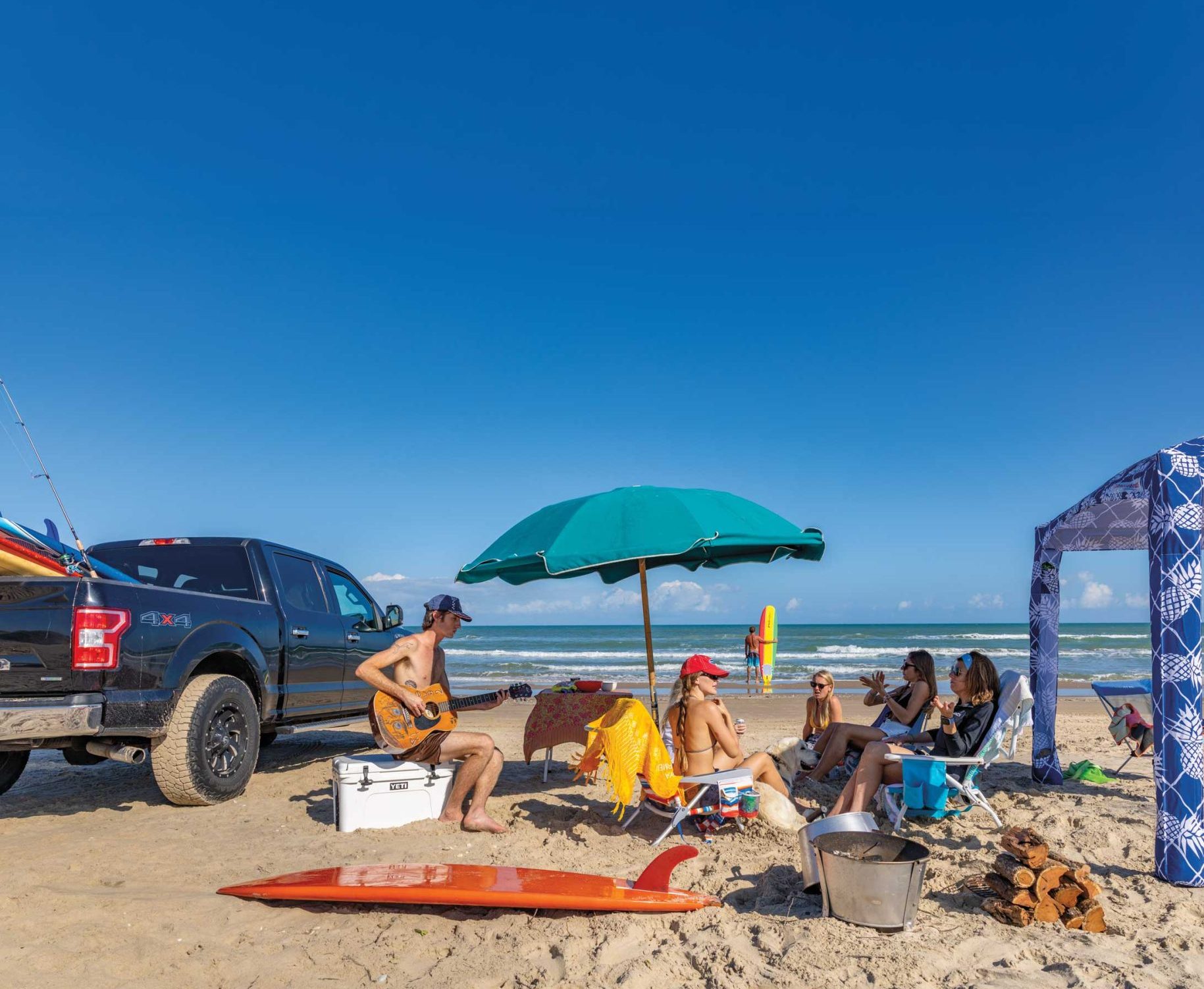 Several people in bathing suits lounge in beach chairs under an umbrella and next to a truck in front of the ocean