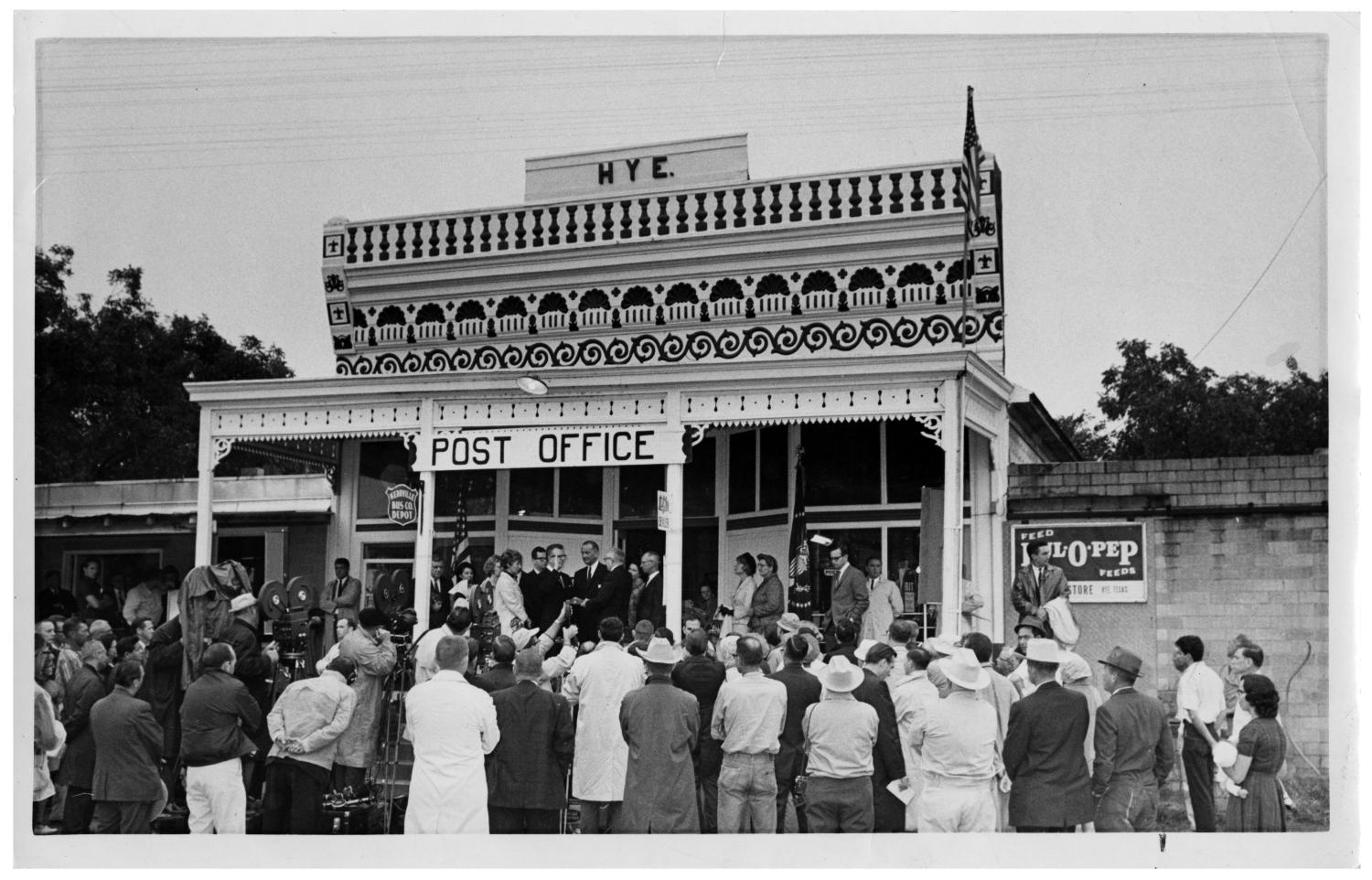 A black-and-white photograph of President Lyndon Johnson standing amid a large crowd of onlookers in front of a white building that says HYE at the top and has a sign that says POST OFFICE
