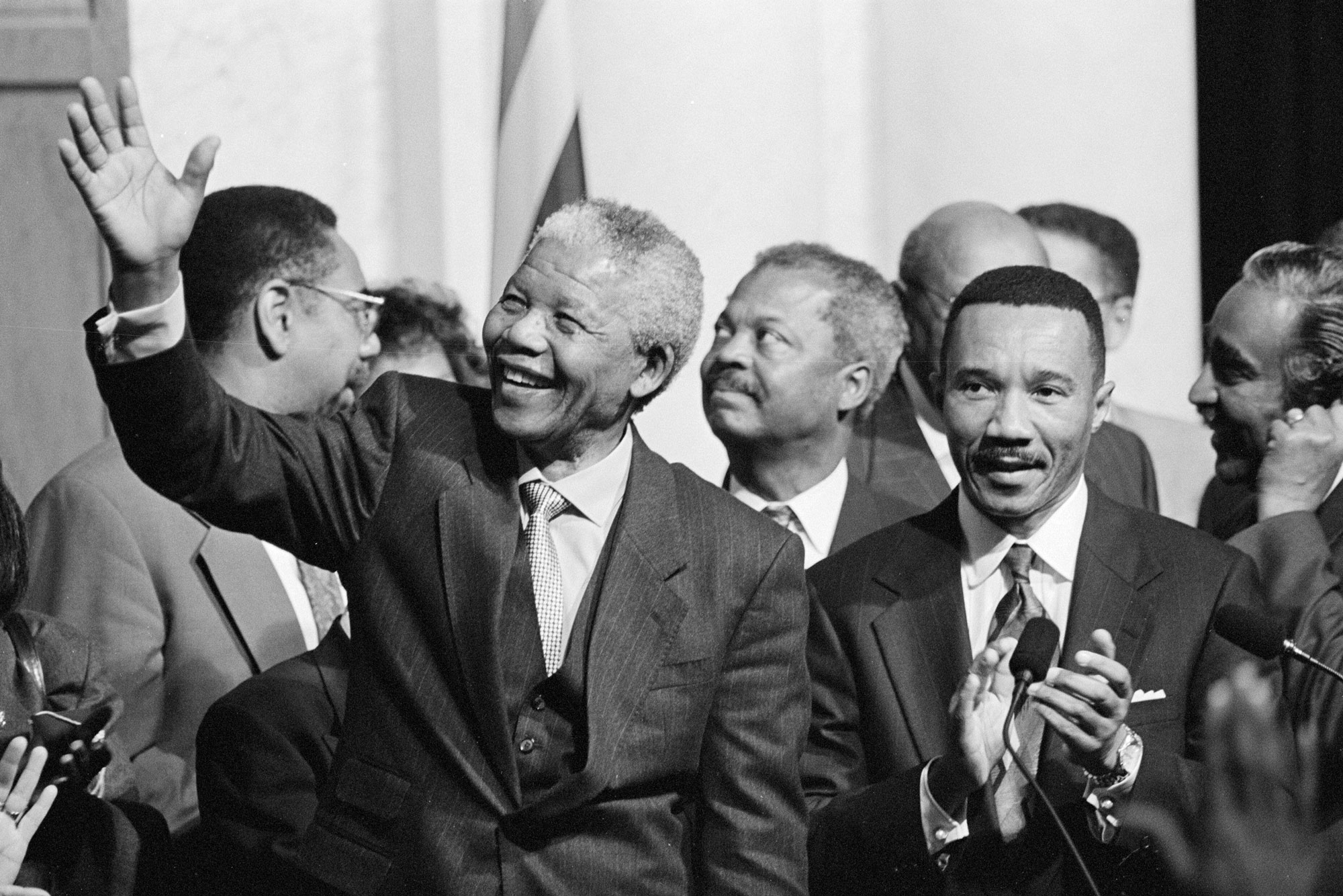 A group of black men in suits stand in front of microphone and a flag. An older man in front smiles and waves