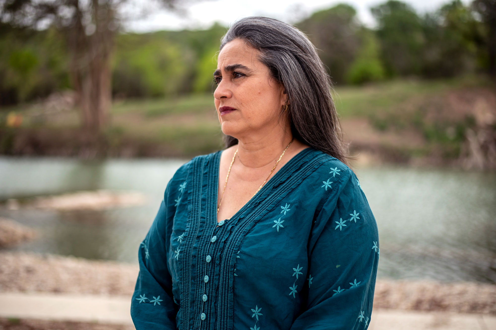 A woman with dark grey hair in a teal top stares solemnly off into the distance in front of a river