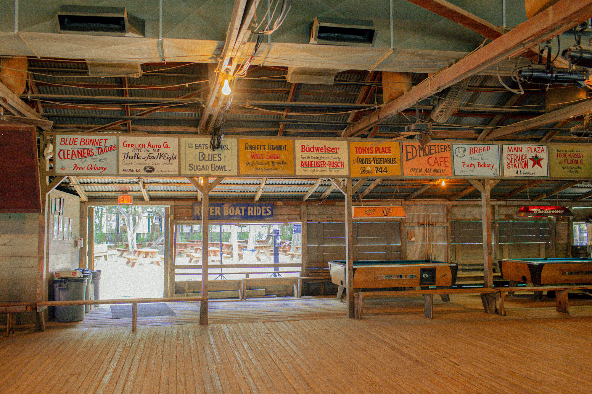 A dance hall with wooden floors and rafters, two pool tables, and many vintage advertisements