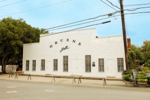 A white building with black text that reads Gruene Hall with plants, trees, and shrubbery next to it