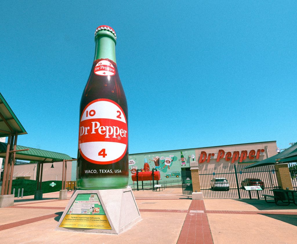 A giant statue of a glass soda bottle that reads "Dr Pepper" on the label stands in a courtyard. Behind it is a building that has a sign that reads "Dr pepper"
