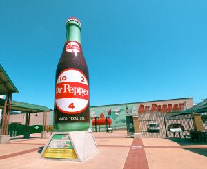 A giant statue of a glass soda bottle that reads "Dr Pepper" on the label stands in a courtyard. Behind it is a building that has a sign that reads "Dr pepper"
