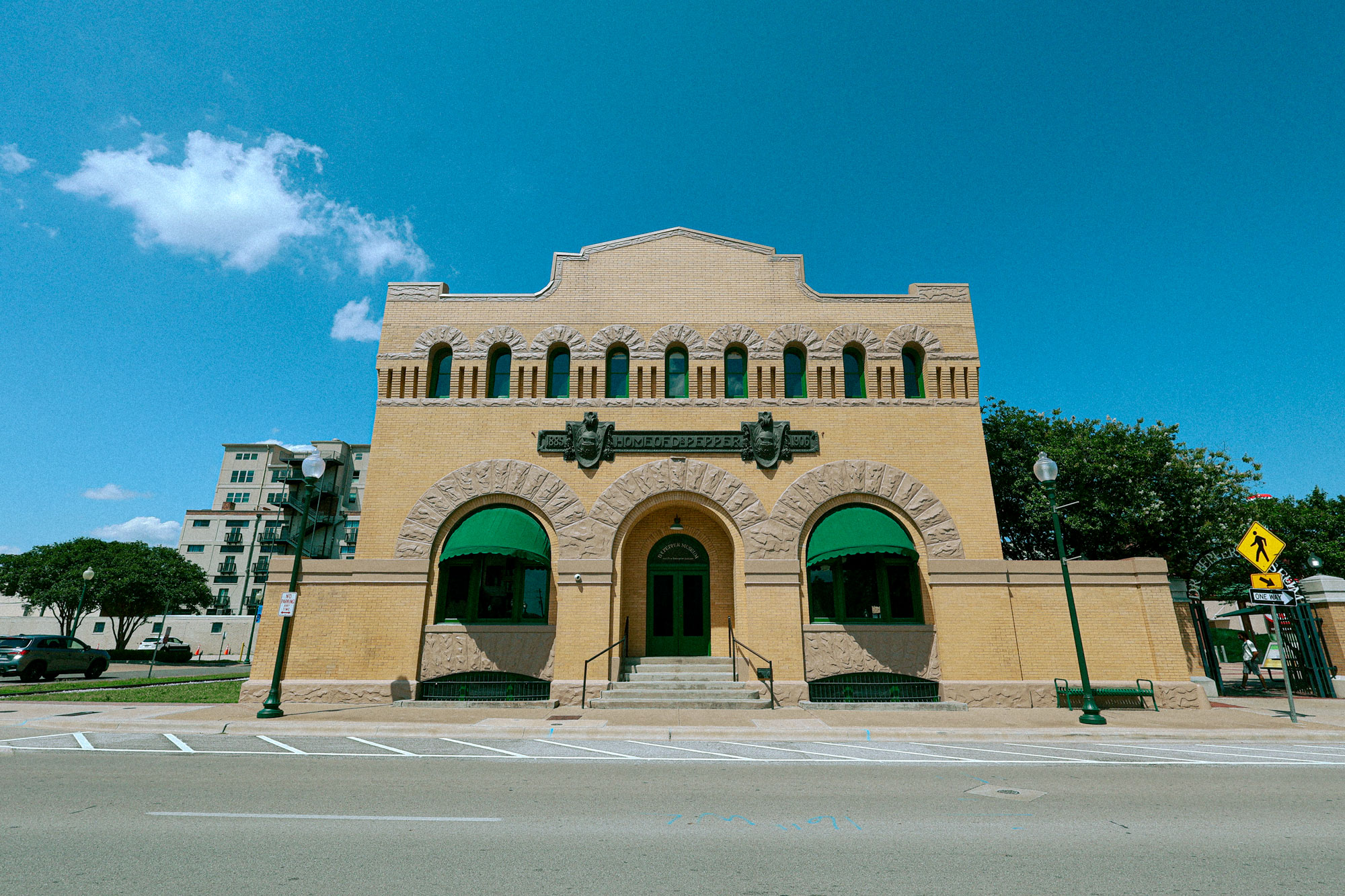 A beige brick building that resembles a train station with green awnings over the windows. An engraved stone sign reads "Home of Dr Pepper"