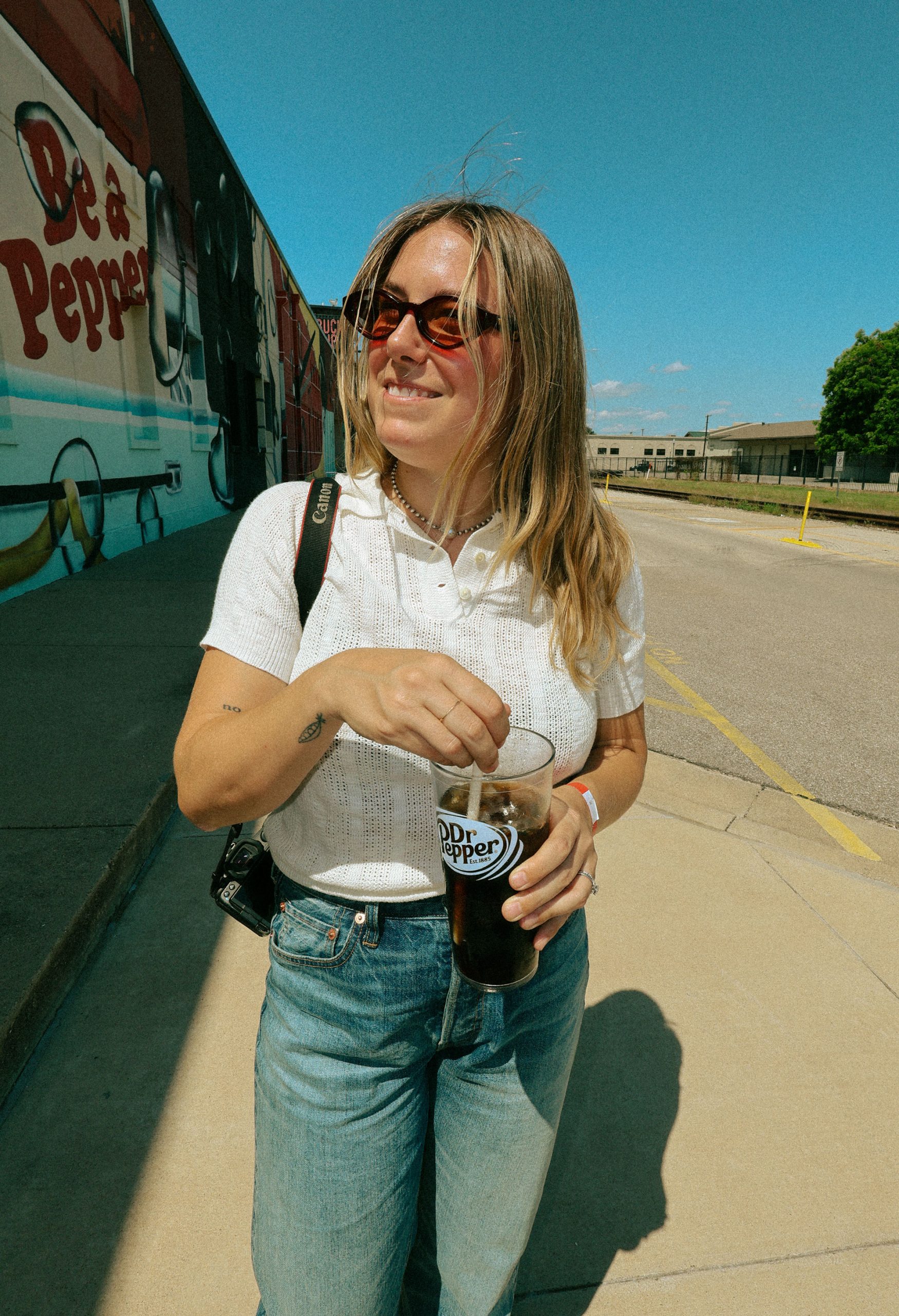 A blonde woman in red sunglasses, a white shirt, and jeans holds a plastic cup full of dark liquid that reads "Dr Pepper" on the side. A mural that reads "Be A Pepper" is behind her.