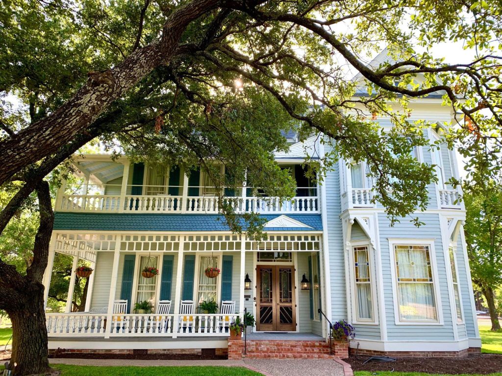 A multi-story baby blue house with darker blue shutters and a brown wood door behind a large tree