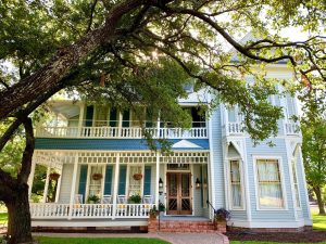 A multi-story baby blue house with darker blue shutters and a brown wood door behind a large tree