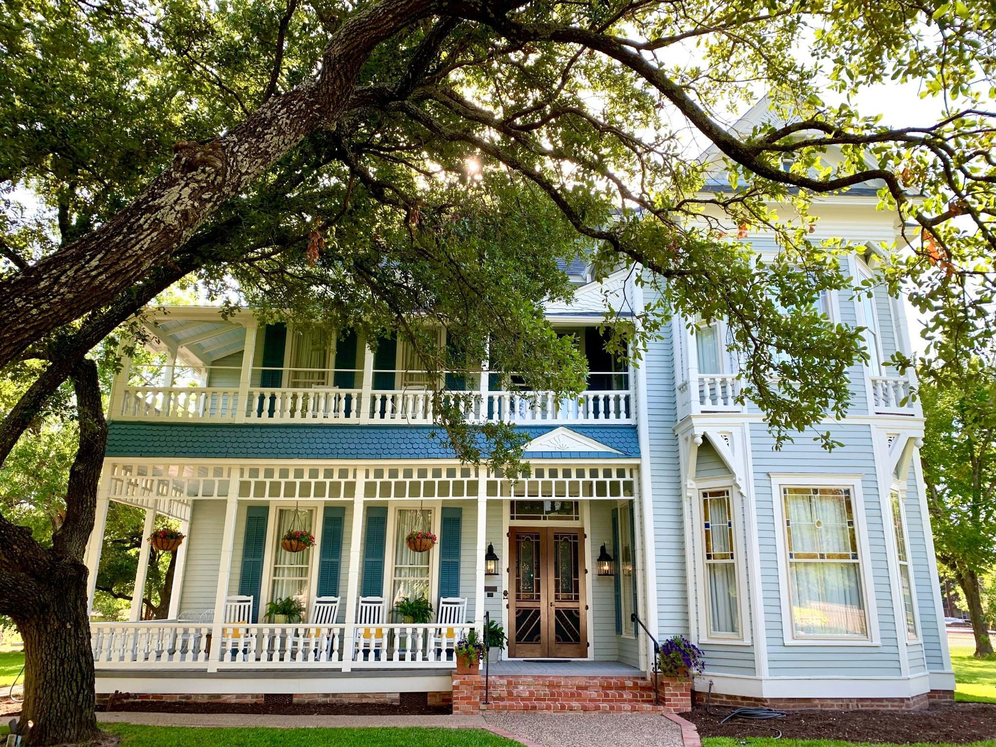 A multi-story baby blue house with darker blue shutters and a brown wood door behind a large tree