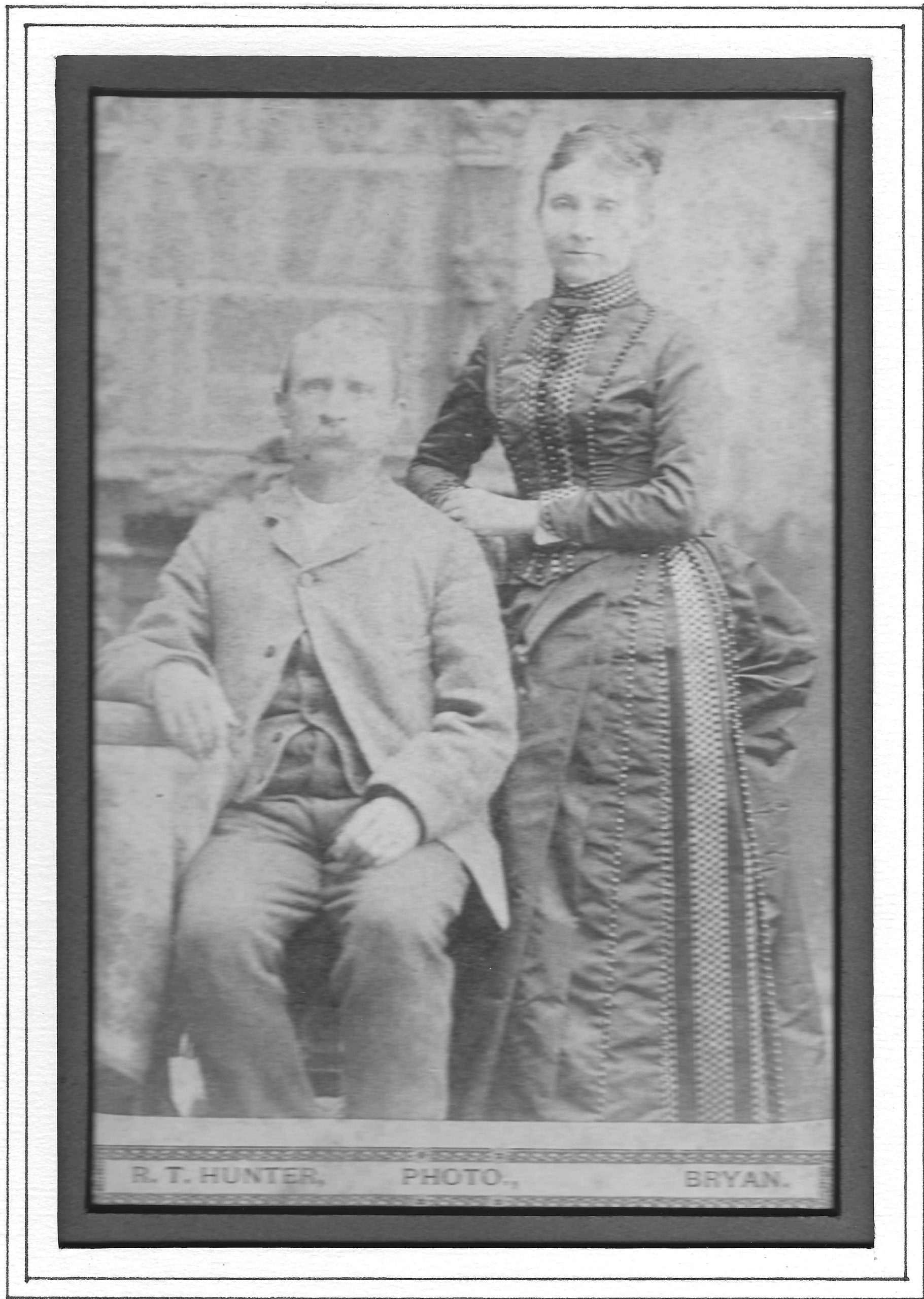 A seated man next to a woman, both in formal dress and with serious looks on their faces
