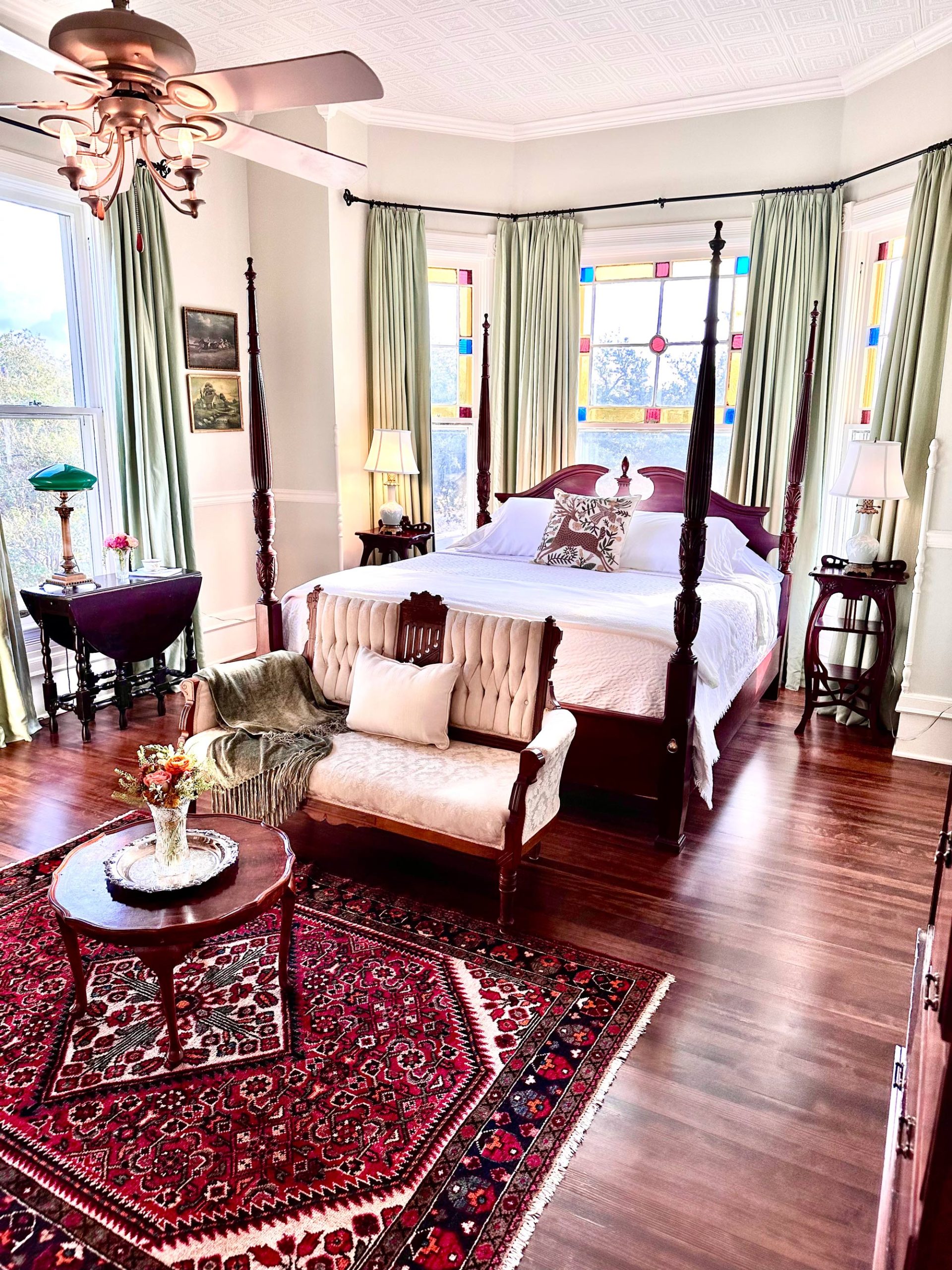 A bedroom featuring a four-post bed with a white comforter, stained glass windows, decorative green and white lamps, a settee, a wooden coffee table, and a red and white rug