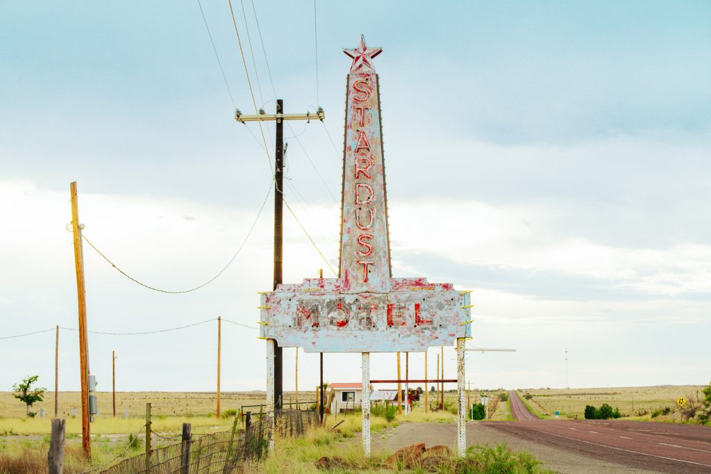 A faded blue sign with a star on top that reads Stardust Motel on the left side of a deserted road
