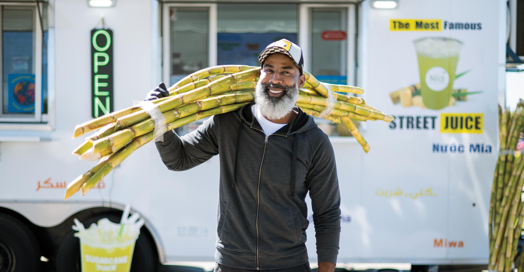 A man with a white beard and a baseball cap on holds a big bundle of green sugarcane on his shoulder