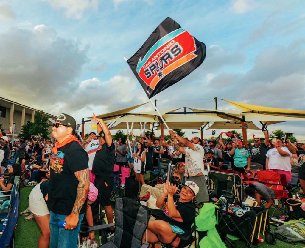 A group of San Antonio Spurs fans in a field and under a large yellow awning, one of whom is waving a large San Antonio Spurs flag
