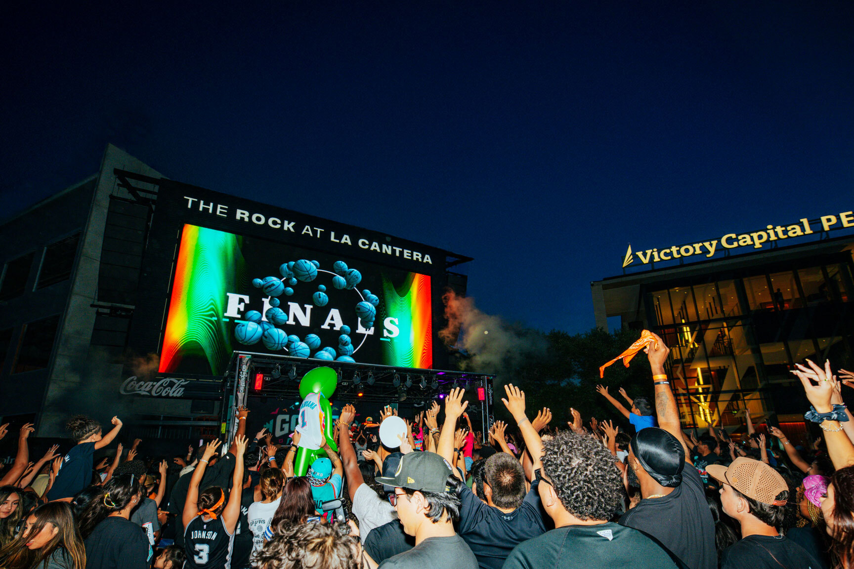 A large group of Spurs fans at night in front of a large television screen that reads FINALS