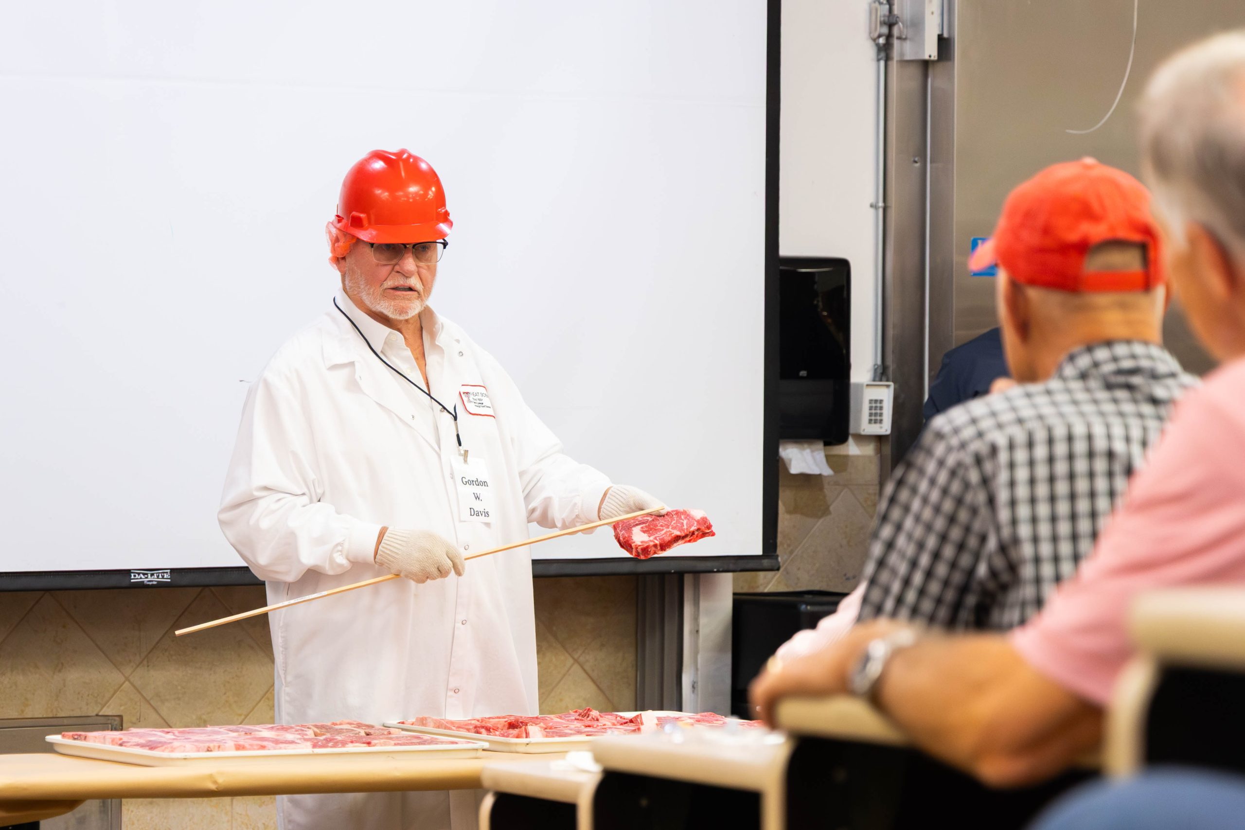 A man in an orange hardhat holds a cut of meat and explains to people seated in front of him about it