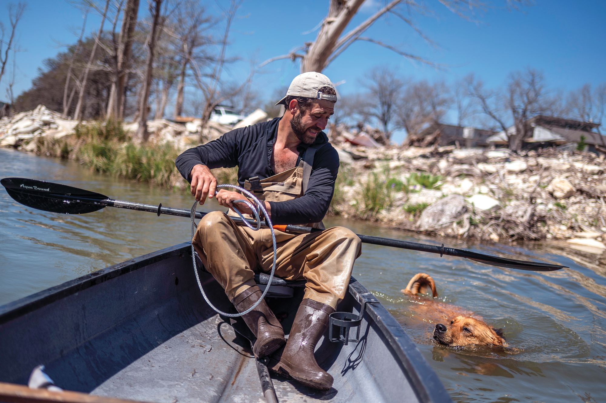 A man in fishing waders sits in a canoe with a paddle while looking at a brown dog swimming in a river next to the watercraft.