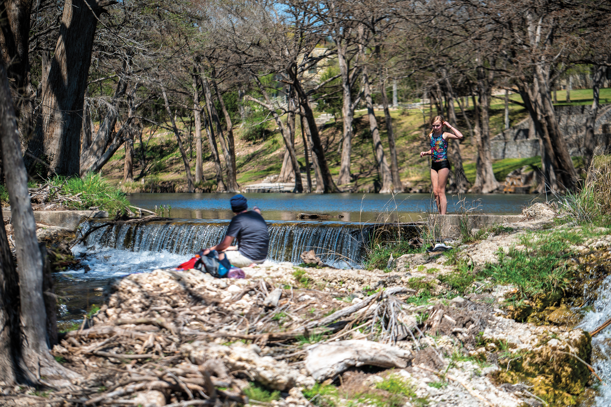 A young girl in a swimsuit stands in a river on a small waterfall. A man sits on rocks while talking to her