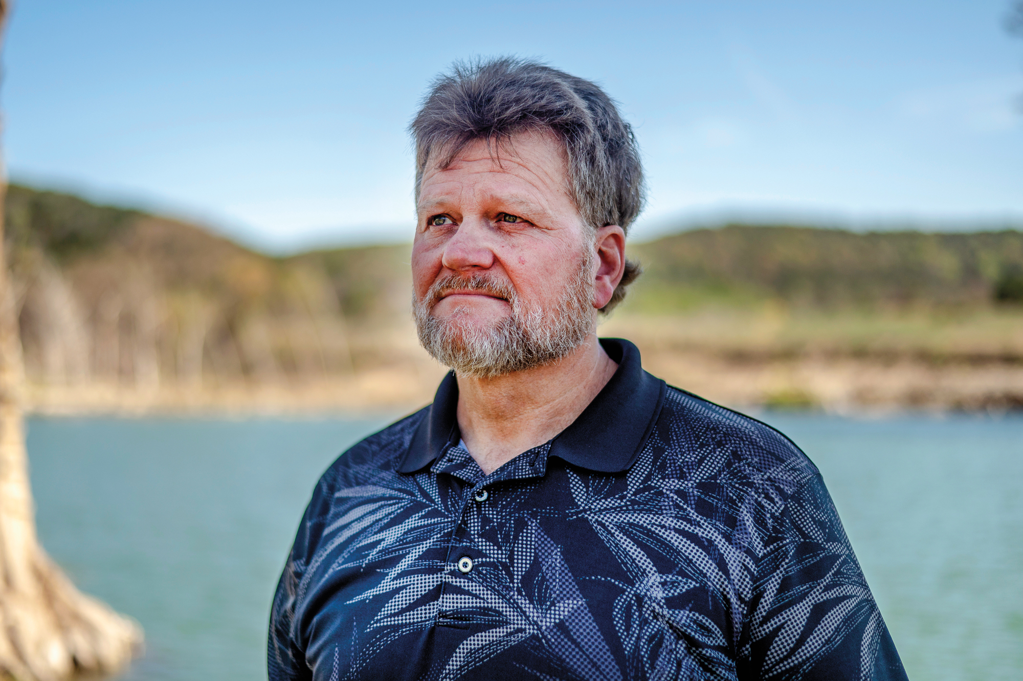 A man in a blue polo poses outside during the day in front of a river with a solemn expression