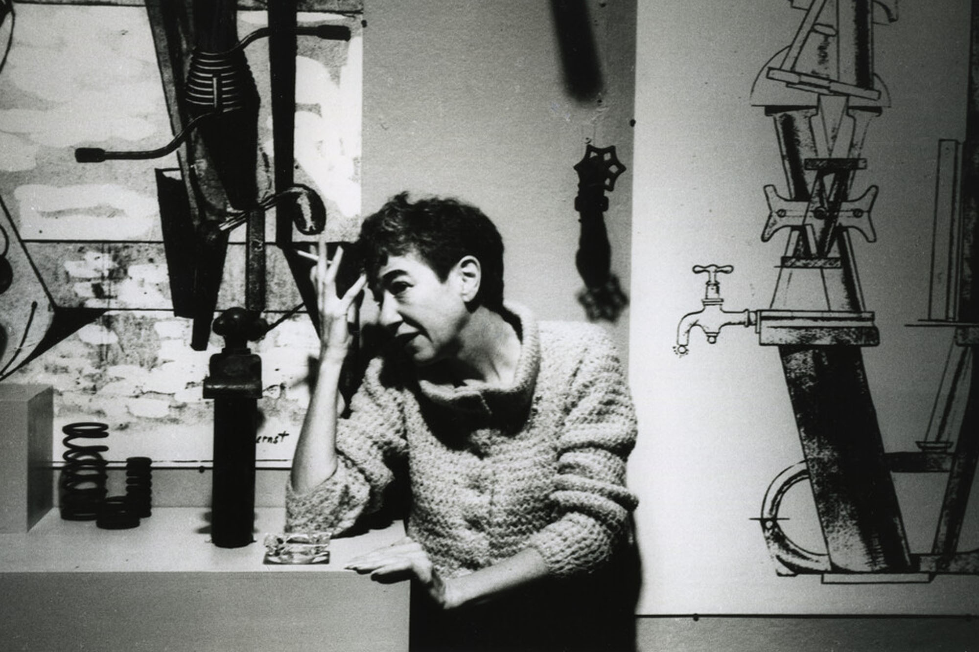A woman wearing a sweater holding a cigarette to her forehead in front of a white wall with pipework painted on.