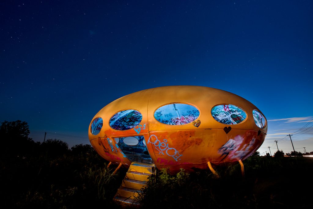 A weathered, bright orange saucer-shaped house with a graffitied interior in a grassy lot at night.