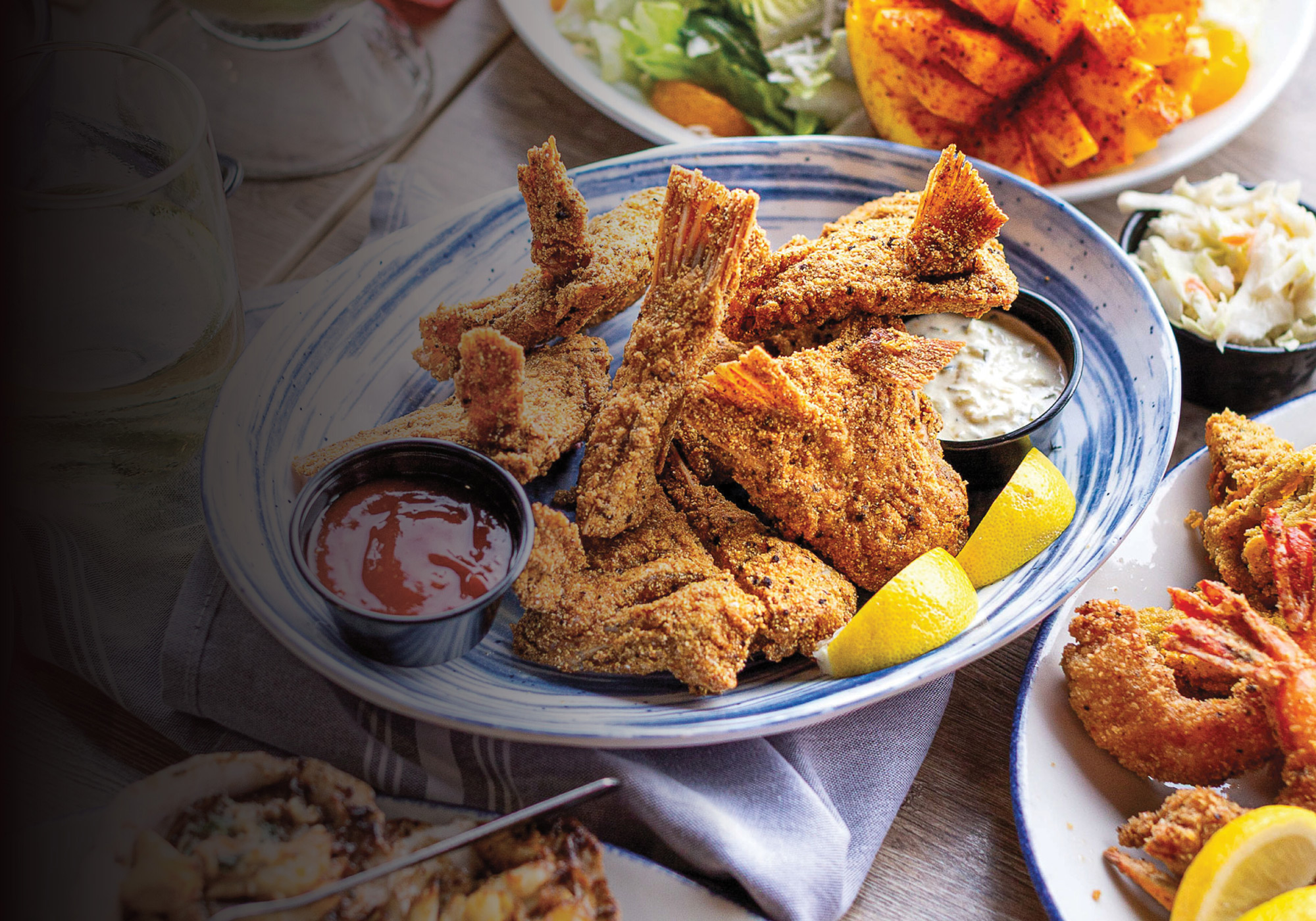 A blue and white shallow dish filled with fried seafood, two dipping sauces, and lemon wedges next to a glass of white wine