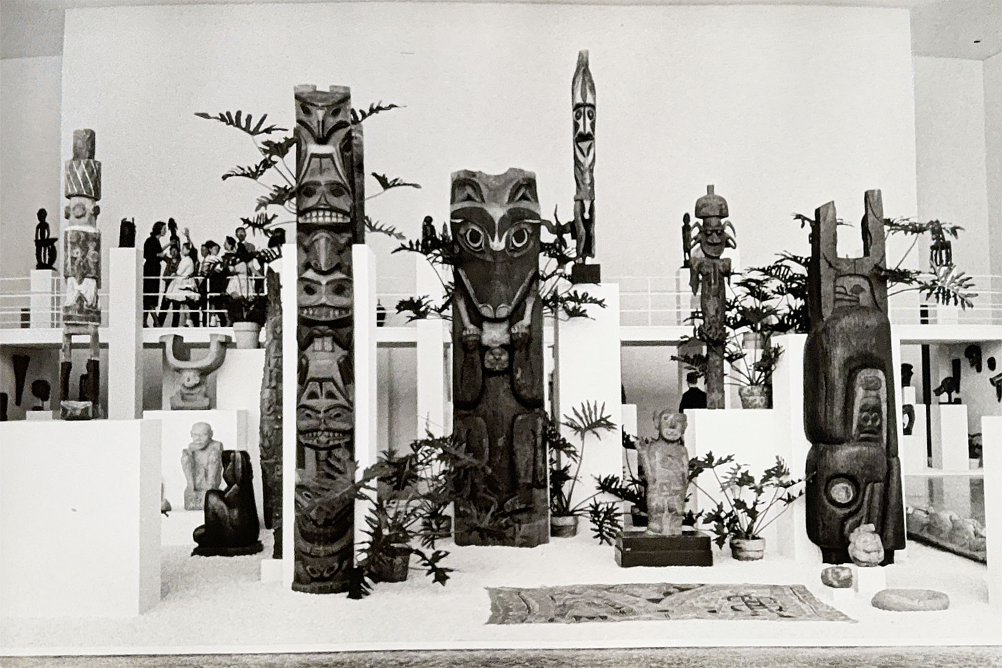 An arrangement of wood totem poles, stone figures, and plants in a large, white gallery space with walkways.
