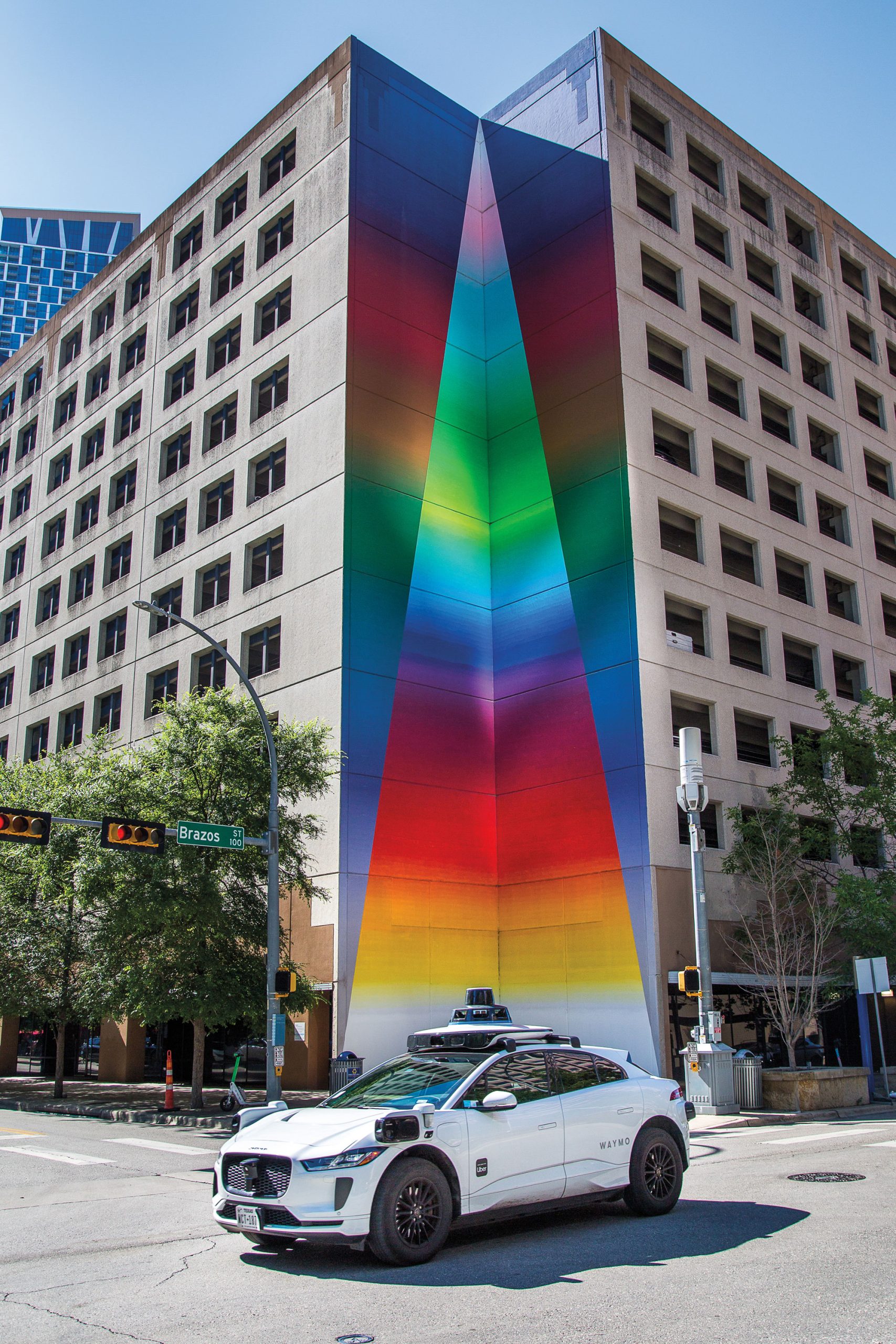 A Waymo drives through an intersection in front of a mural of a multi-colored triangle.