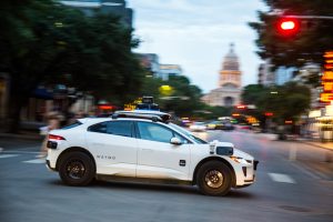 A white hatchback vehicle with cameras on the top and sides attached drives down a street with a fuzzy background of an old Capitol building and streetlight