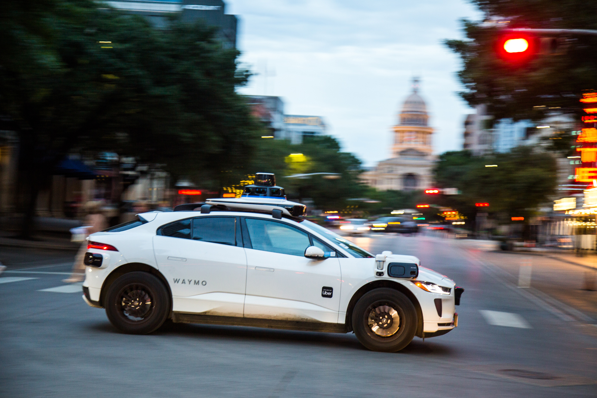 A white hatchback vehicle with cameras on the top and sides attached drives down a street with a fuzzy background of an old Capitol building and streetlight