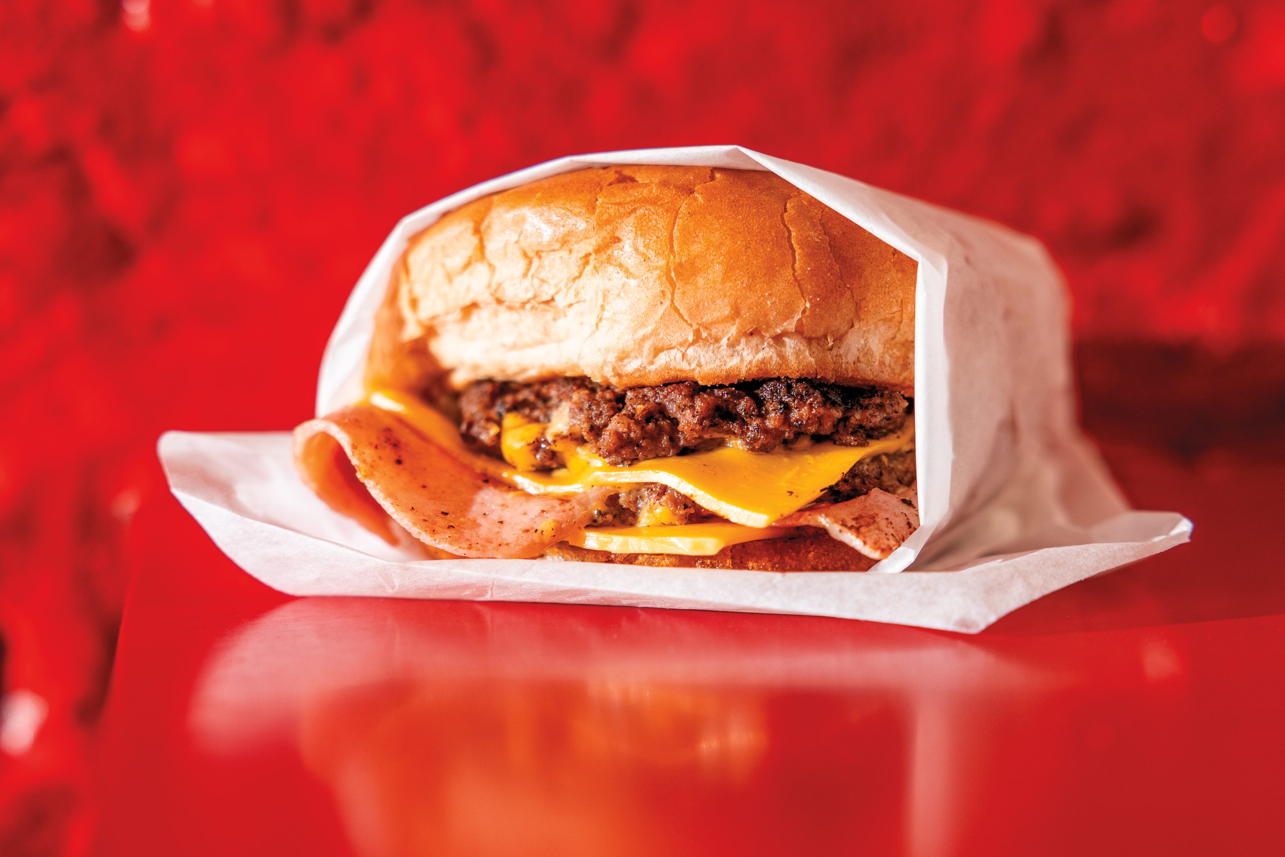 A cheesburger in a white paper wrap against a red wall and table