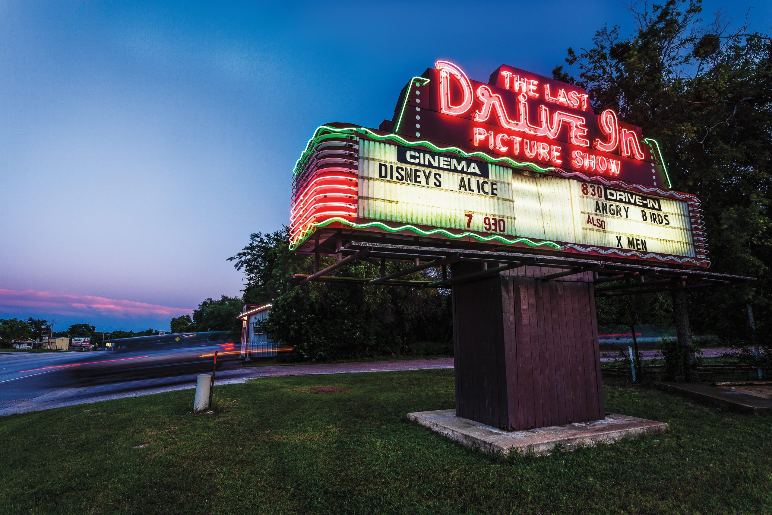 A lit-up neon movie marquee on the side of the road reads "The Last Drive-In Picture Show" backdropped by a purple sunset.
