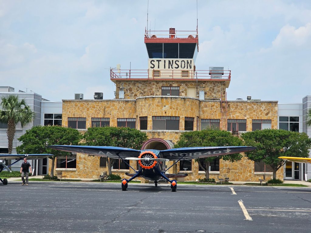A small blue and orange airplane in front of a stone municipal airport with the word Stinson visible on the control tower