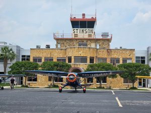 A small blue and orange airplane in front of a stone municipal airport with the word Stinson visible on the control tower