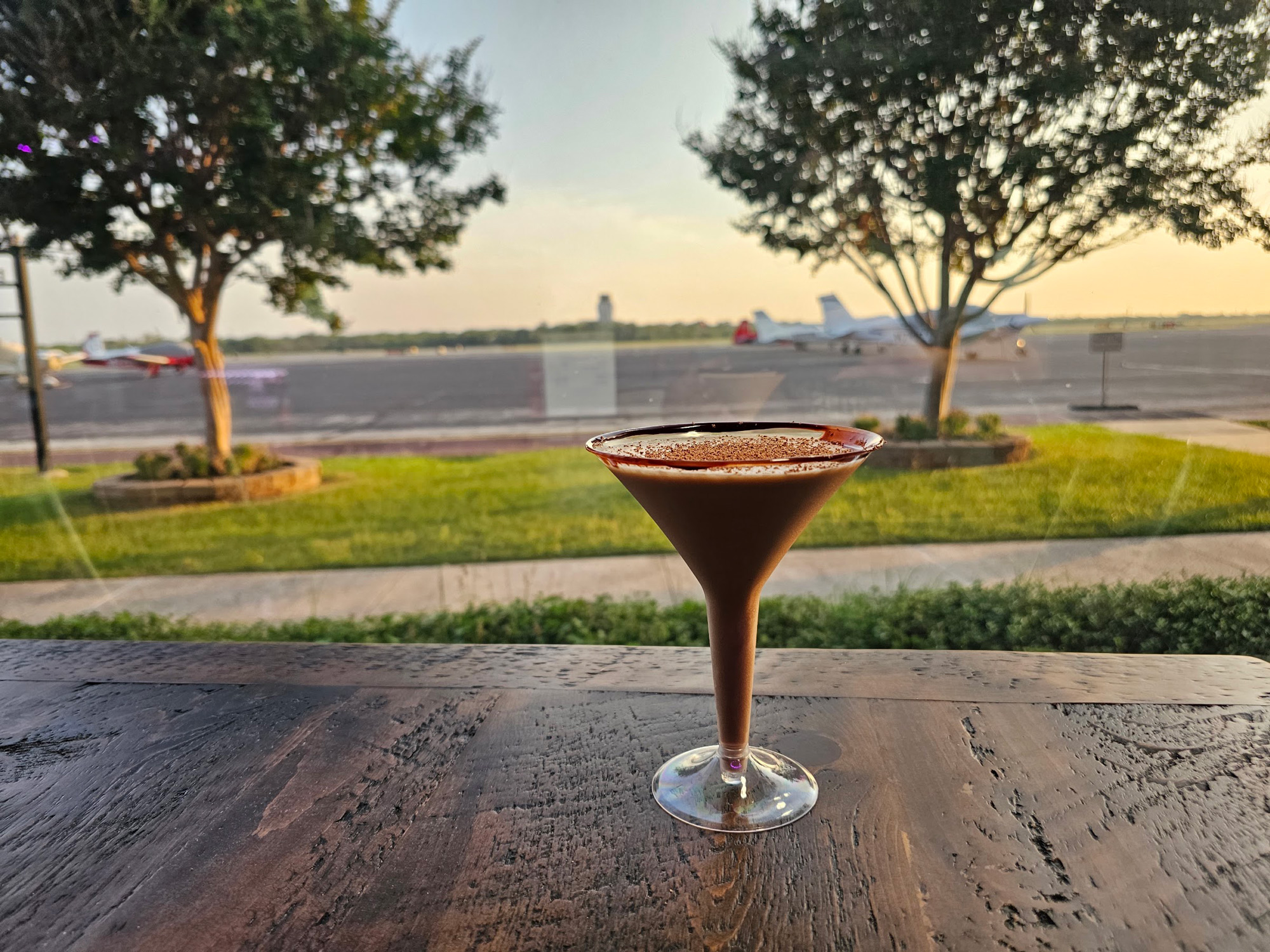 A brown cocktail in a cocktail glass on a wooden table in front of a window with planes on a tarmac visible outside