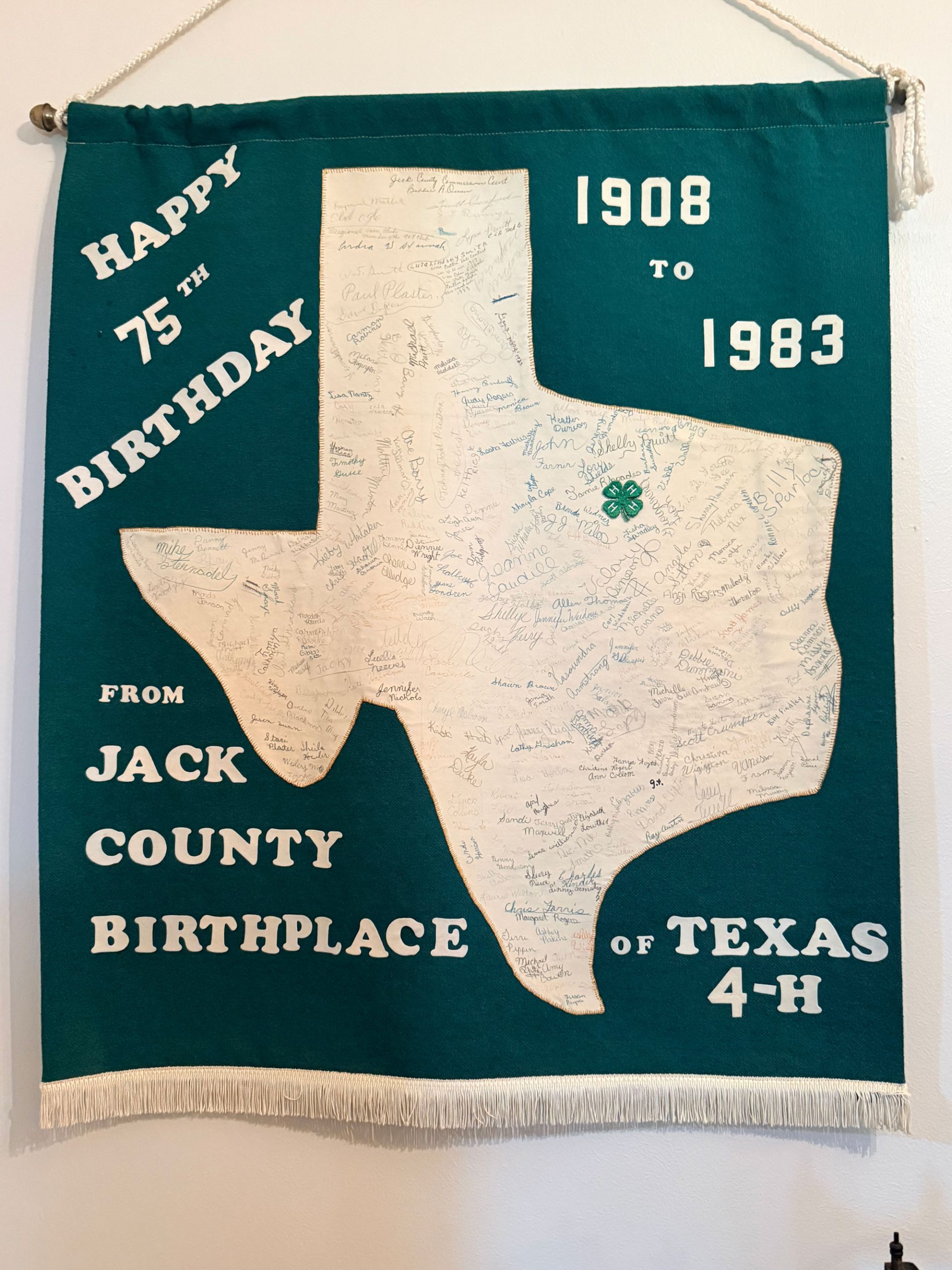 A dark green vertical banner with a white Texas on it that reads "Happy 75th Birthday, 1908 to 1983. From Jack County Birthplace of Texas 4-H."