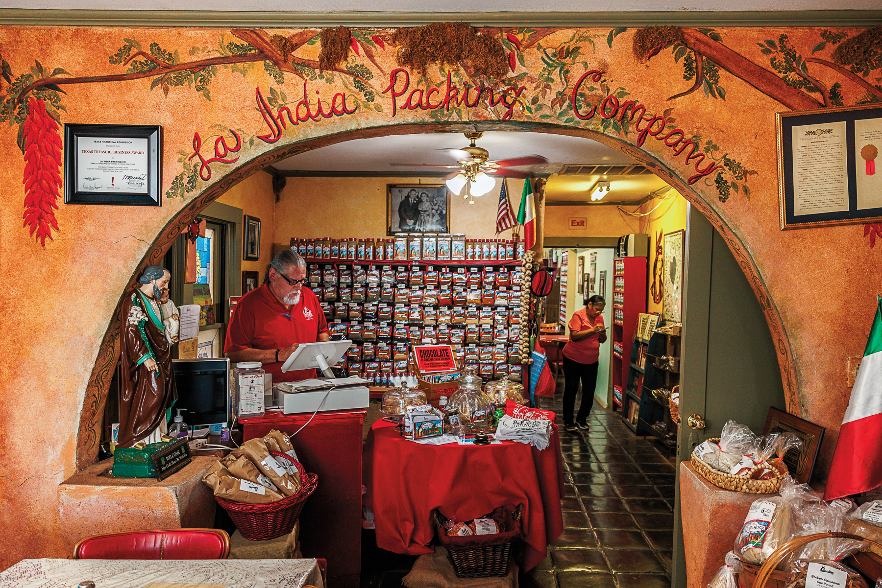 A man works a register inside a small orange-hued building filled with racks of spices for sale