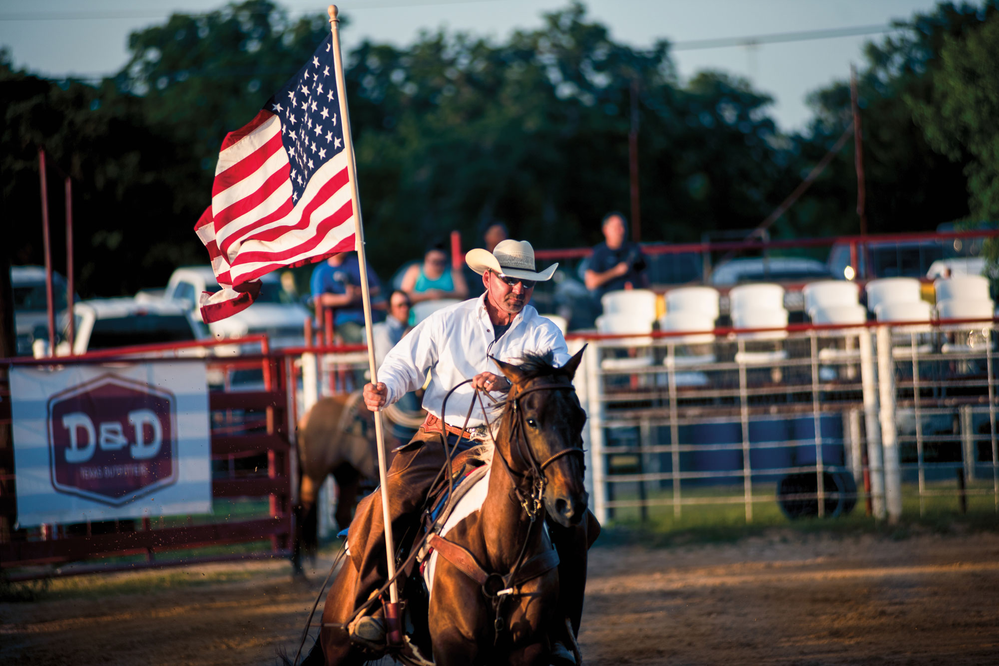 A man in a white cowboy hat and shirt holds an American flag while riding a brown horse around a rodeo ring.
