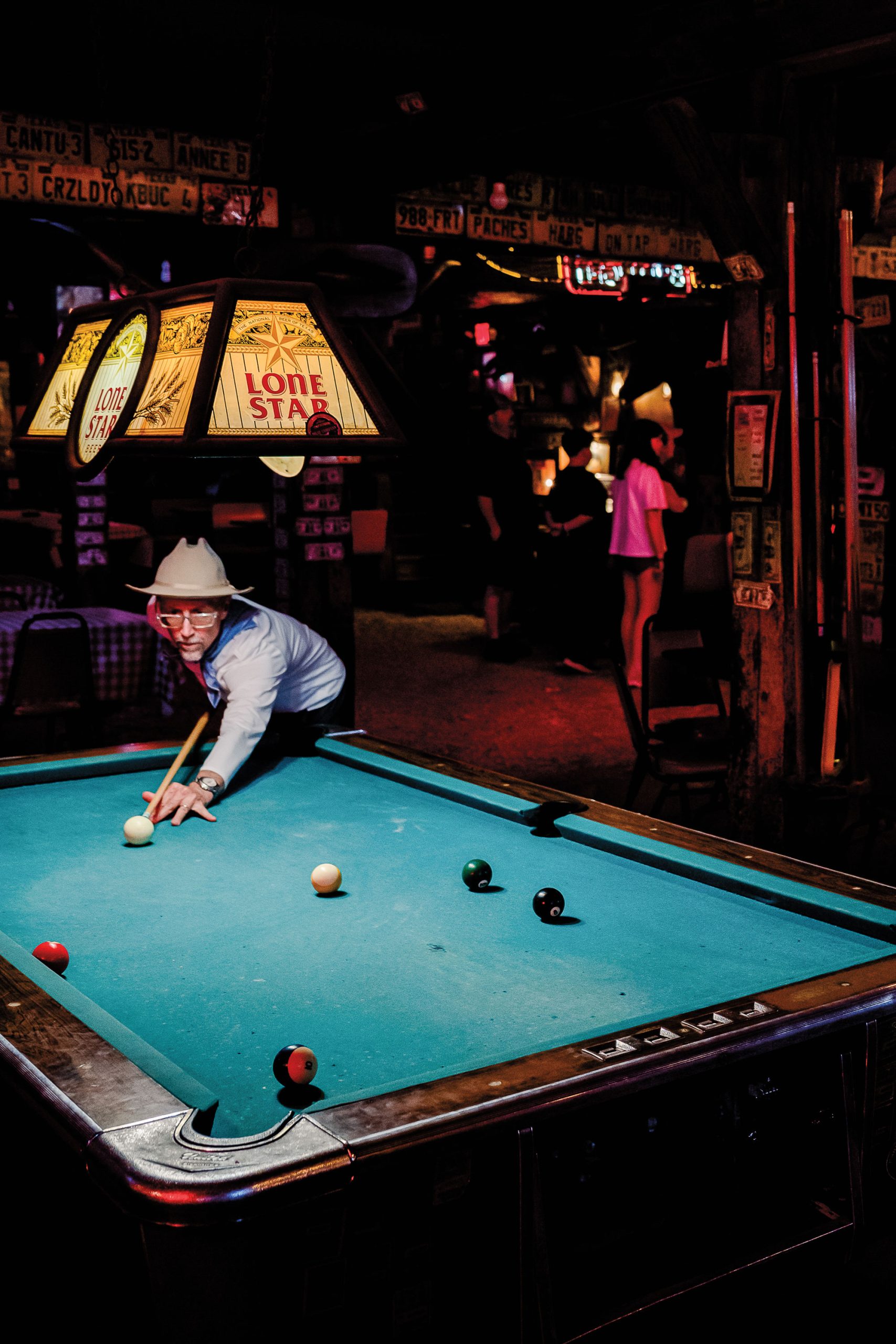 A man in glasses and a long sleeve shirt plays pool underneath a Lone Star light fixture in a dimly lit bar.