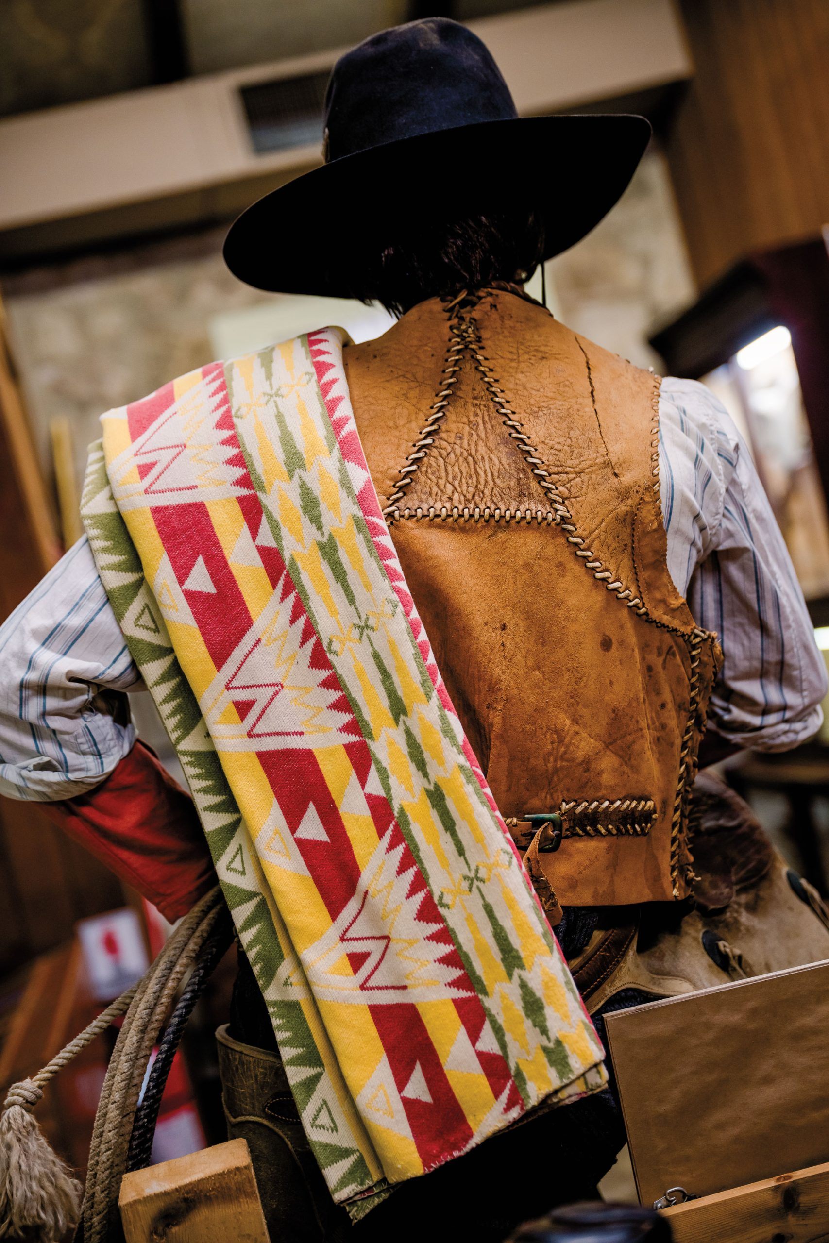 A man in a leather vest and black cowboy hat turns away with a multicolor, patterned blanket over his shoulder.