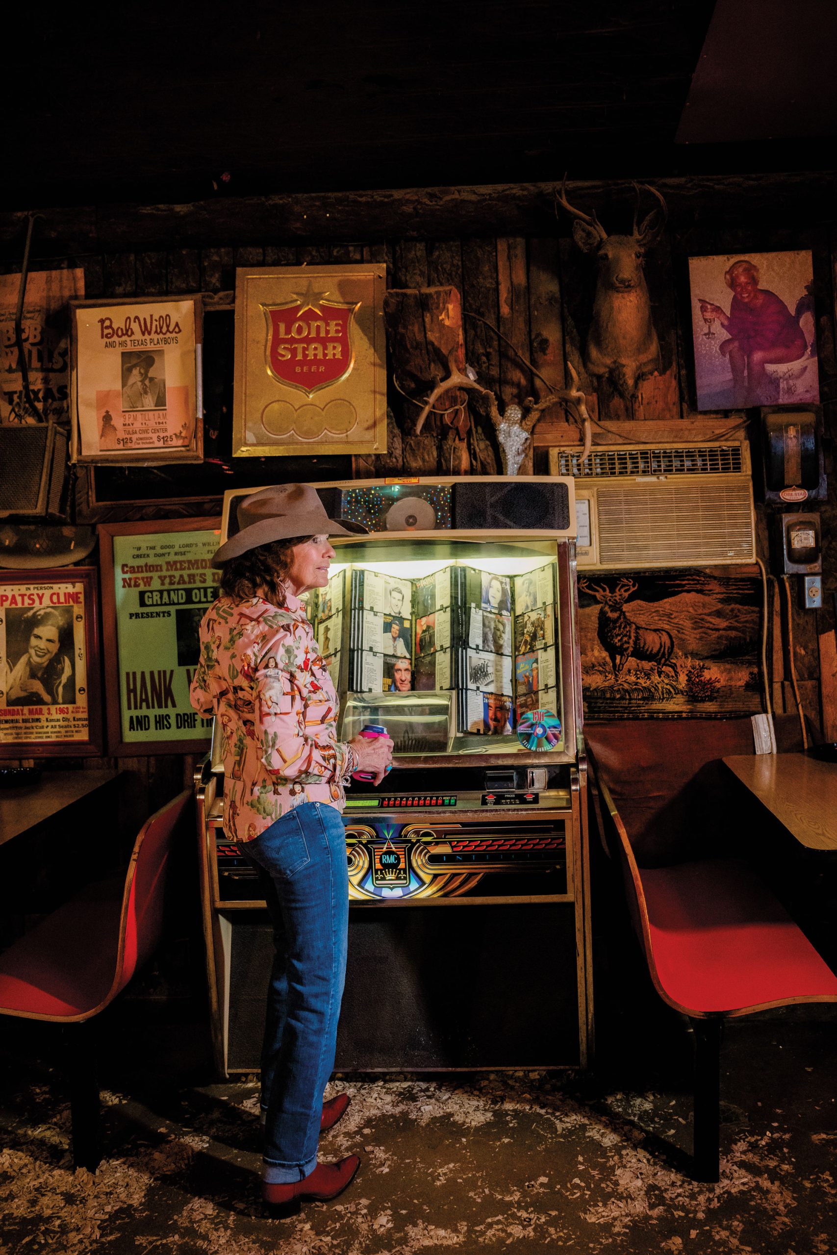 A woman in a cowboy hat, pink shirt, and denim jeans stands in front of a jukebox in a bar.