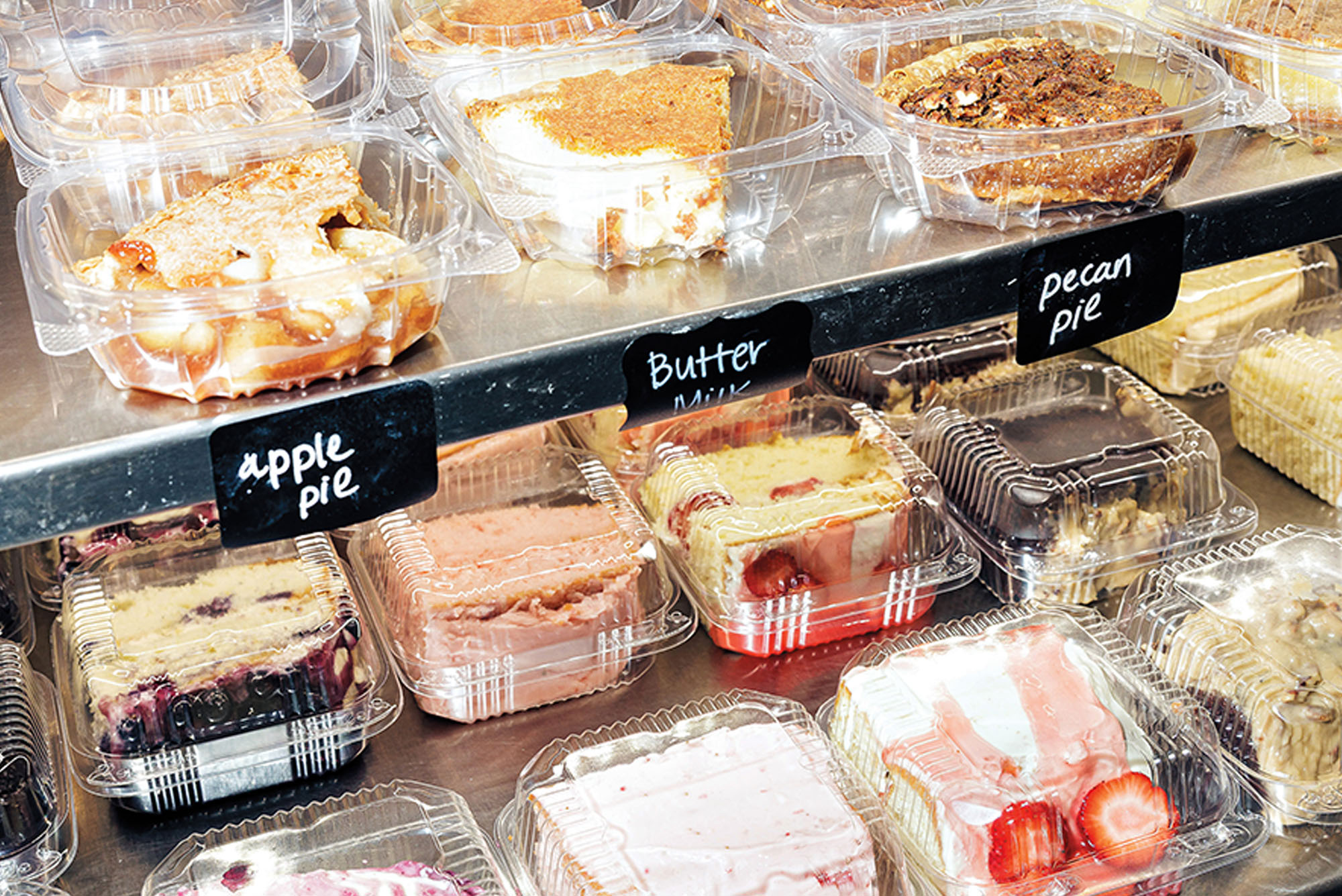 An array of various cakes in clear plastic boxes on labeled shelves.