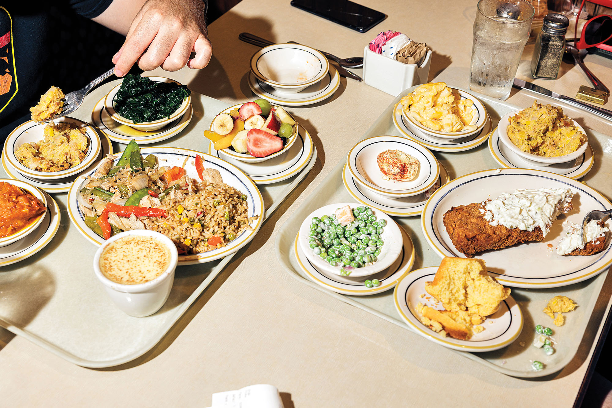 Two filled trays of cafeteria food, with a hand holding a fork over the leftmost one.