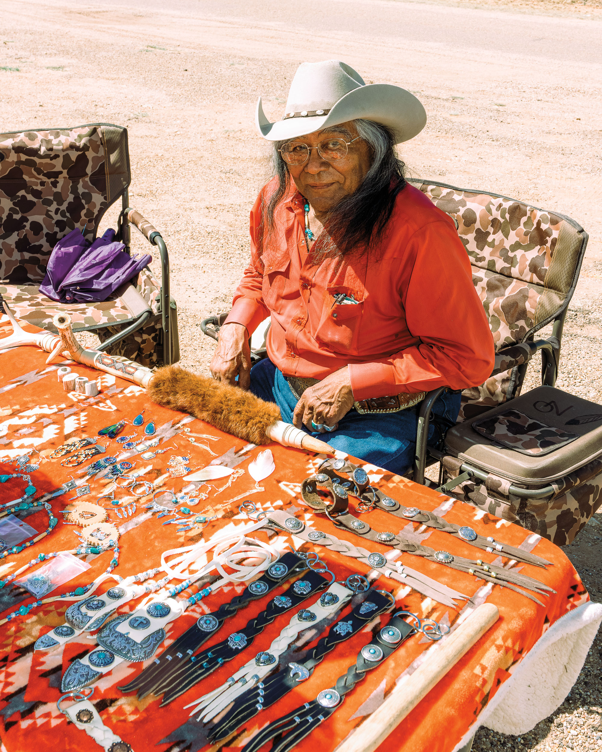 A woman in an orange button down top and a cowboy hat sits in a lawn chair in front of a table featuring a spread of handmade jewlery