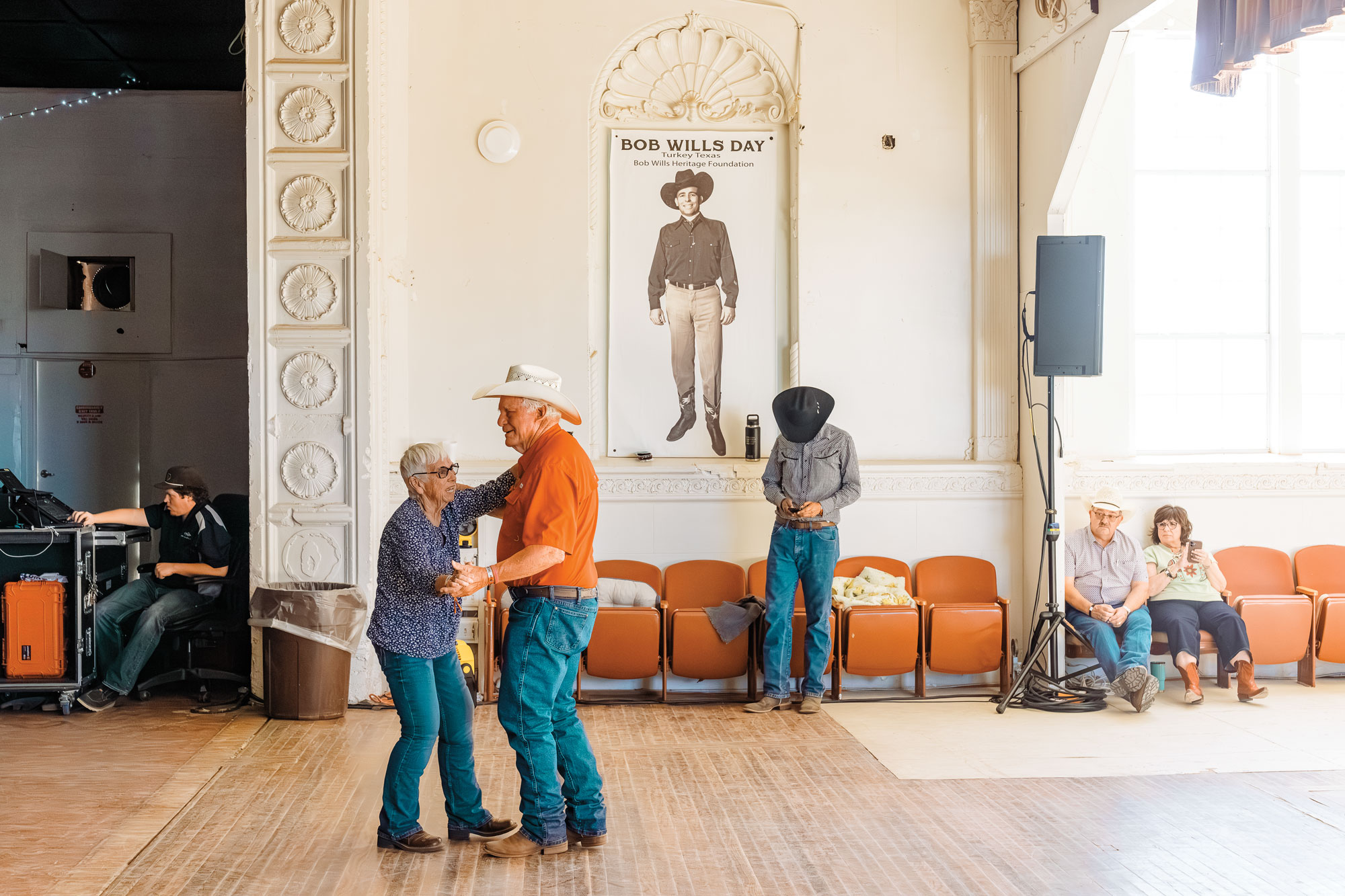 An older couple in Western wear dances in a room with onlookers