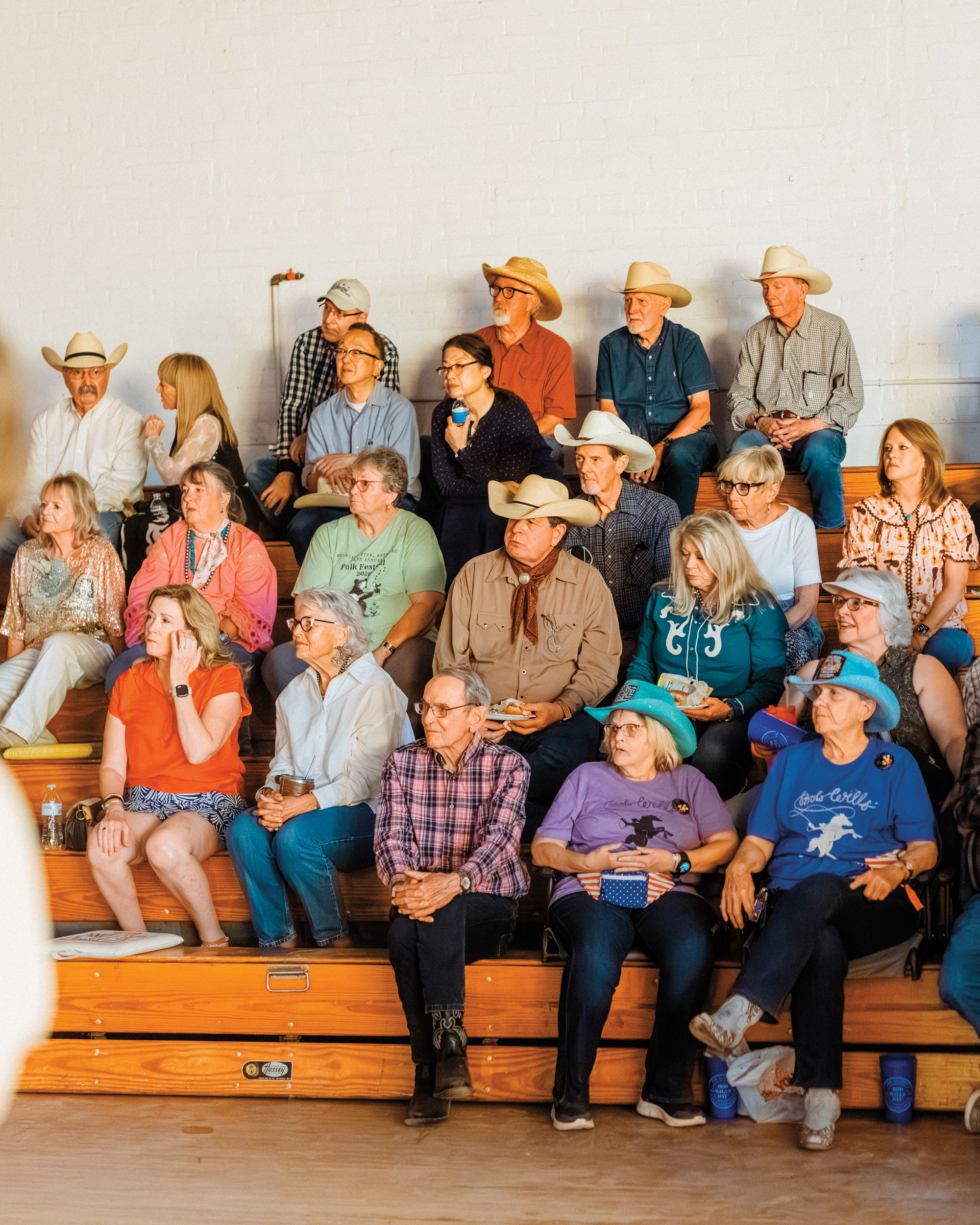 Several people dressed in Western wear sit on wooden bleachers peering off into the distance