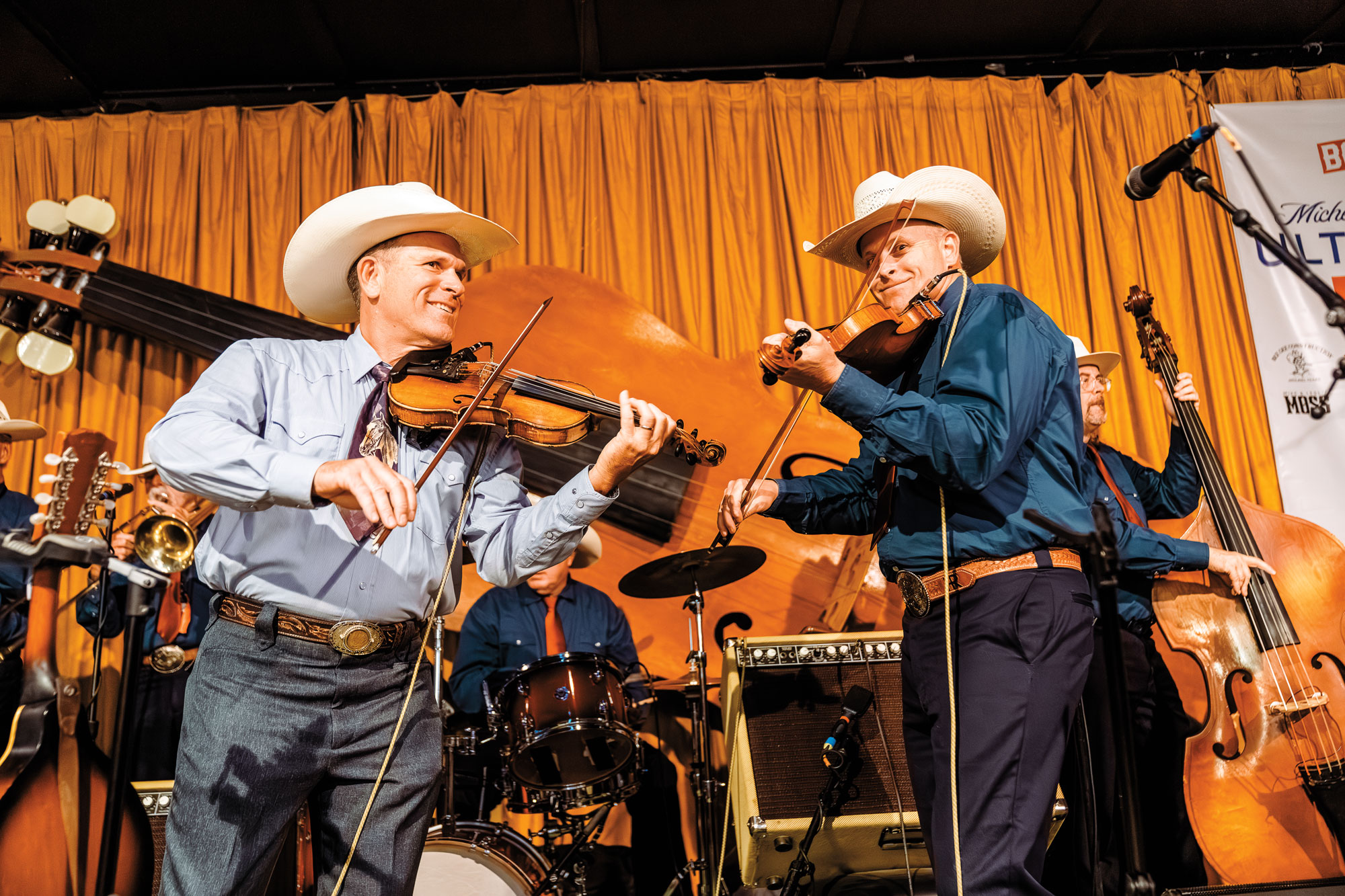 Two men in cowboy hats play fiddles on stage