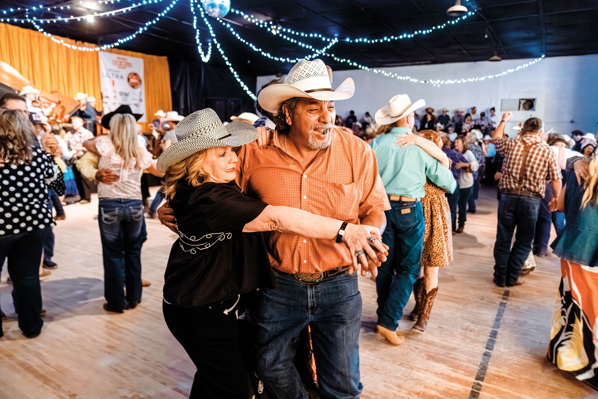 A man and woman in cowboy hats partner dance in a gymnasium decorated with lights and a disco ball filled with other dancers in Western wear