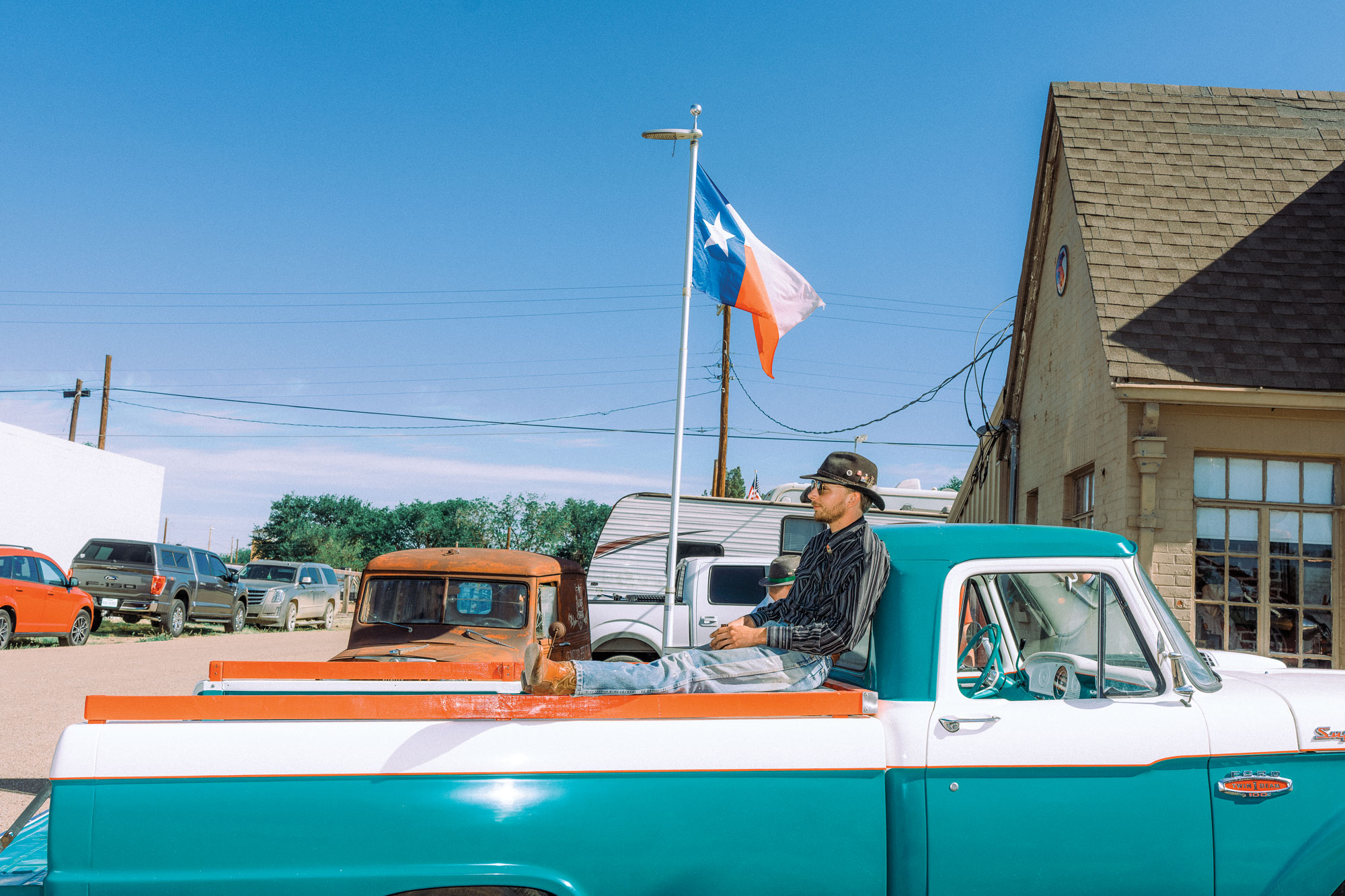 A man in a cowboy hat sits in the bed of vintage blue truck with a Texas flag waving in the background