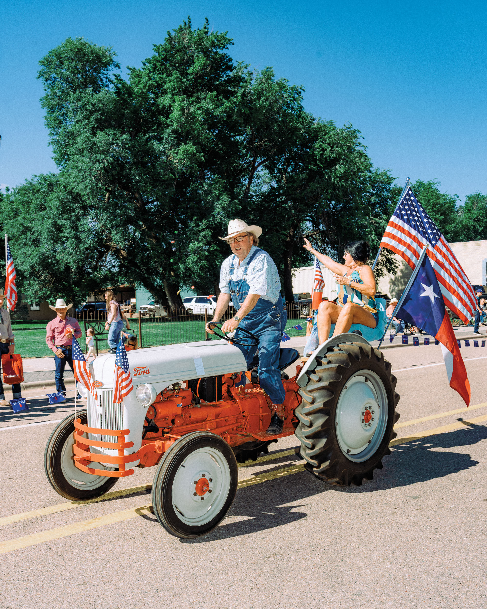 A man in overalls and a cowboy hat rides a vintage tractor through a parade with a American and Texas flags trailing behind him